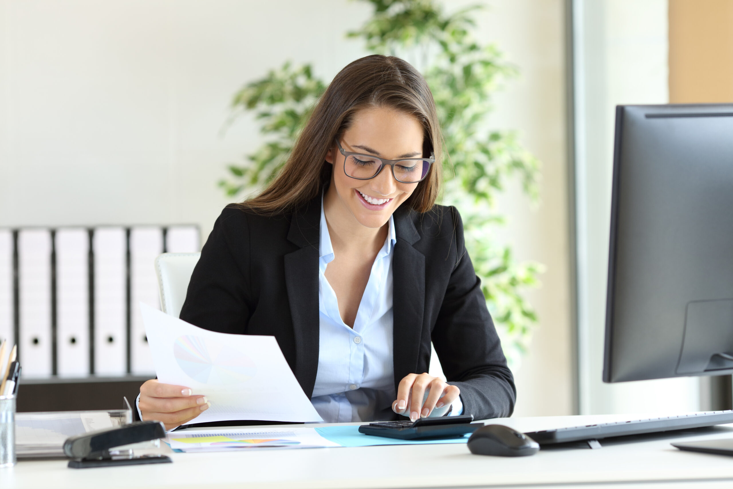 Person smiles while working on a calculator and holding a document. A desktop computer sits in front of them.