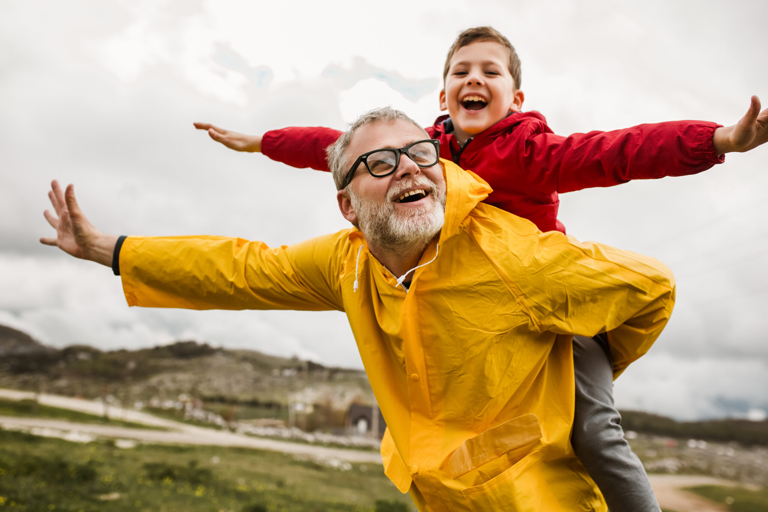 Older person in yellow raincoat smiles while holding a child in a red jacket up on their back. They both appear to be laughing, and are holding their arms out to full wingspan.