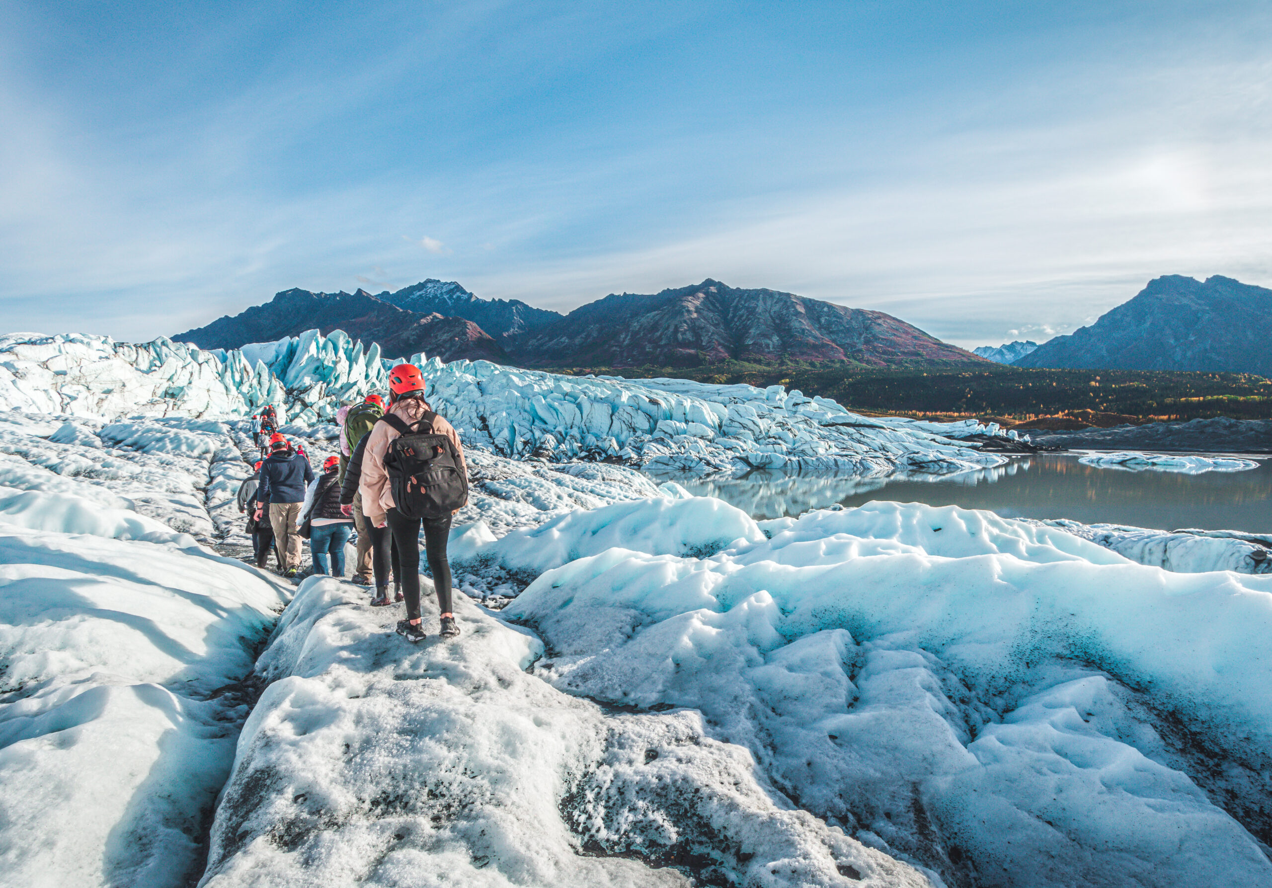 Adventurers in helmets walk away from the camera over an ice field. Mountains can be seen in the background landscape.