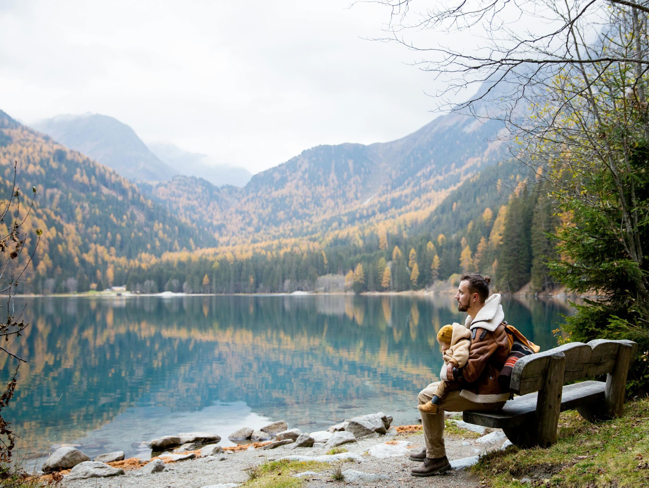 Person sits on a bench while gazing out at a lake and mid-autumn scenery in the mountain landscape behind. They are holding a baby in a yellow hat.