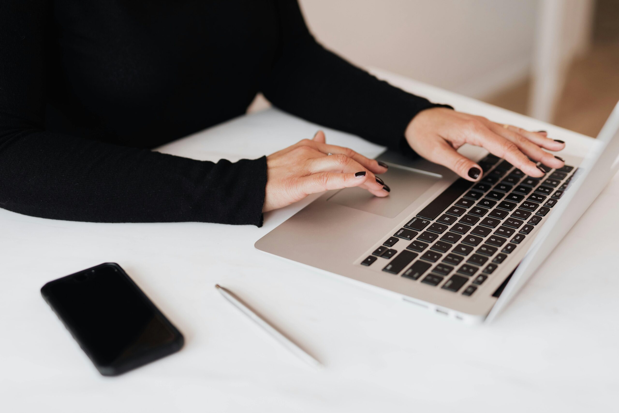 Person with nails painted black types on a laptop. A cell phone and pen sit nearby.