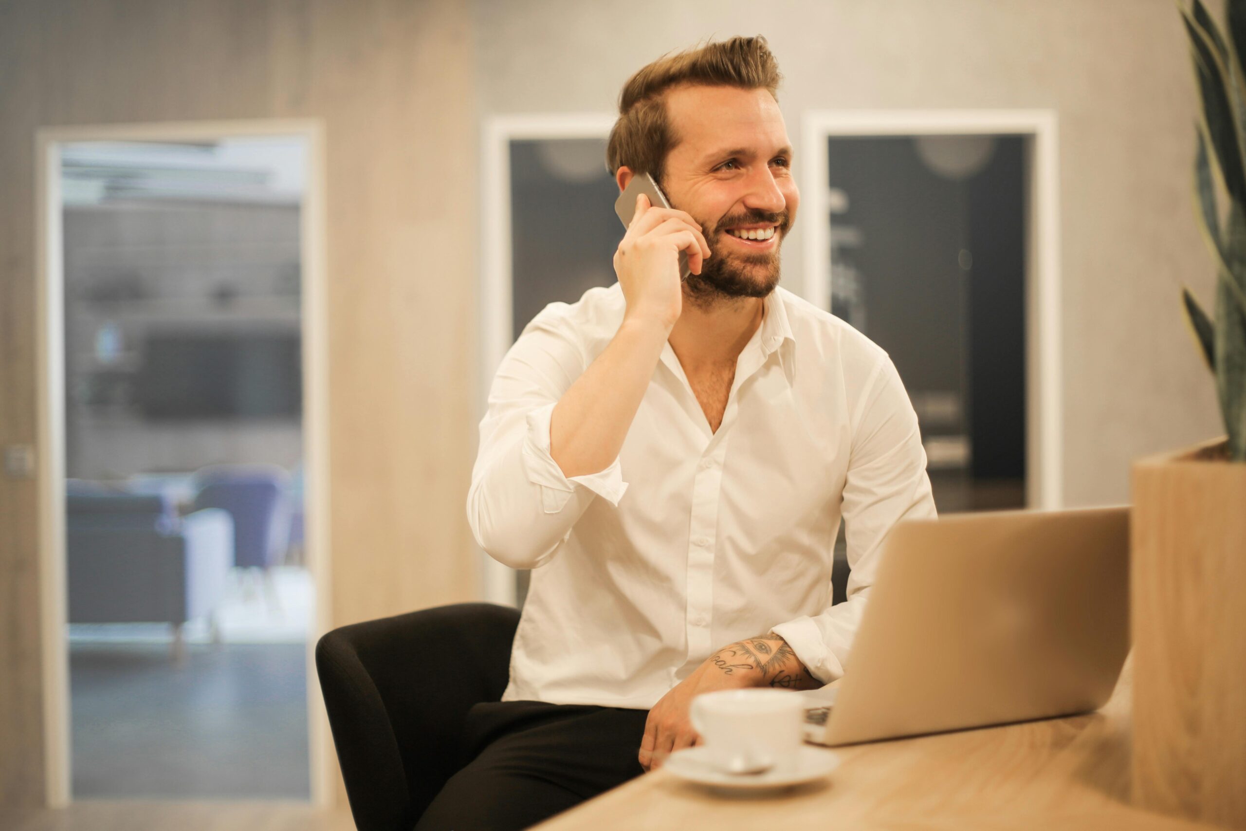 Business dressed individual with coffee and laptop smiles while speaking on the phone.