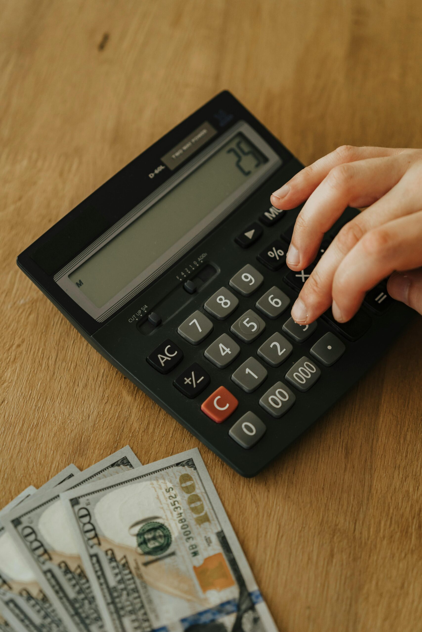 A hand is punching the number '25' into a calculator. A stack of $100 bills can be seen in the lower left corner.